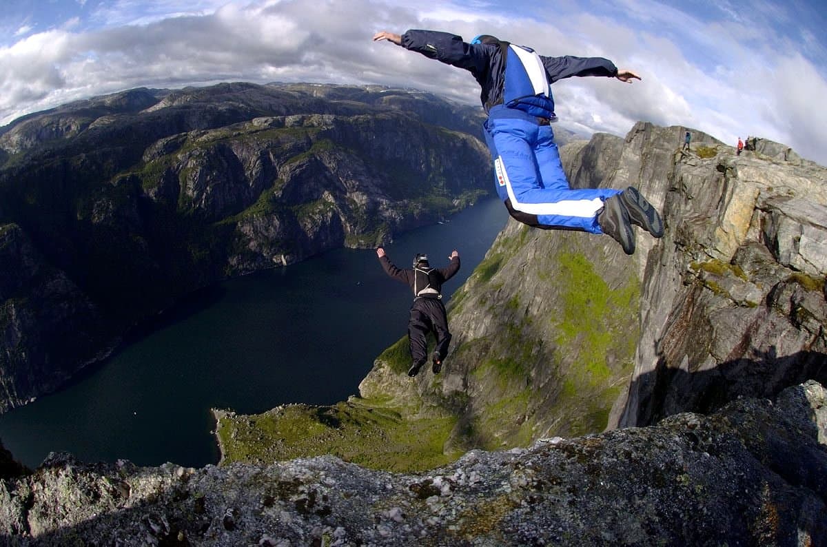 Two people in jumpsuits leave a rocky cliff above a dark stretch of water, with steep mountains. The nearer person wears blue and white, and the other wears black.