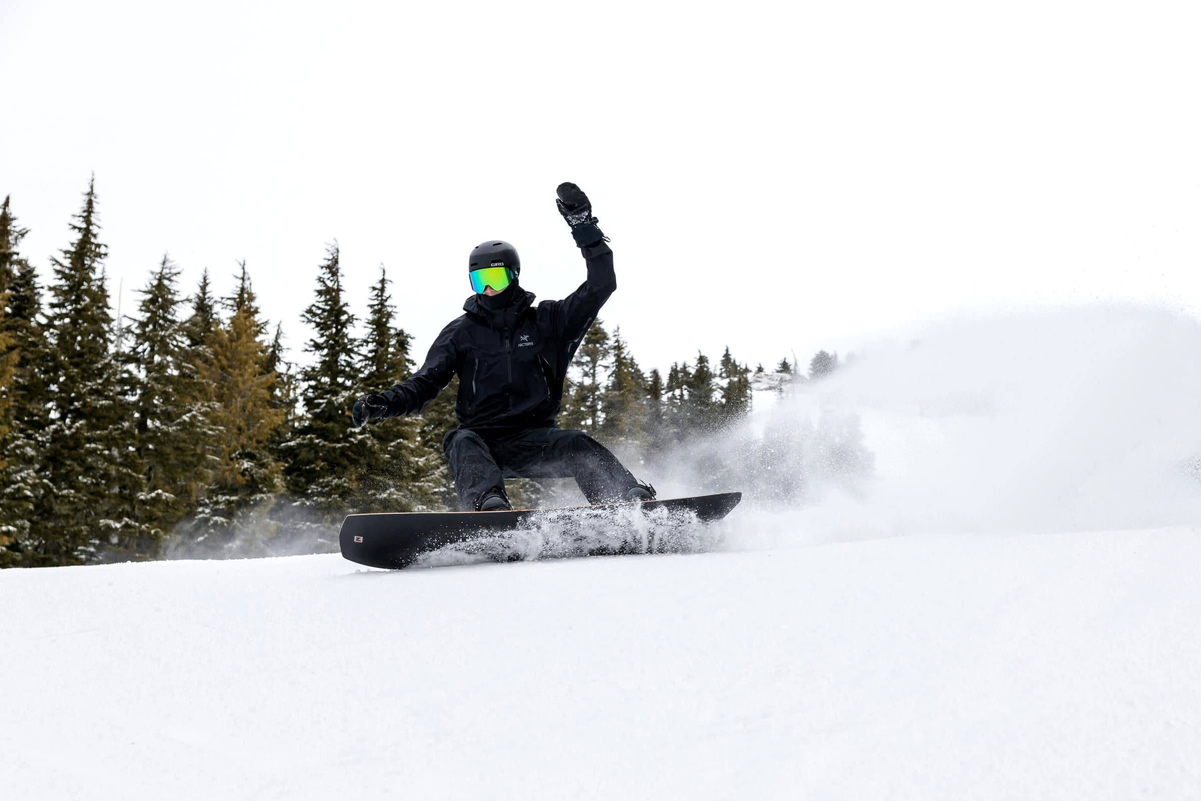 A beginner snowboarder in a helmet and gloves side-slipping on their heel edge down a gentle indoor real-snow slope while an instructor watches