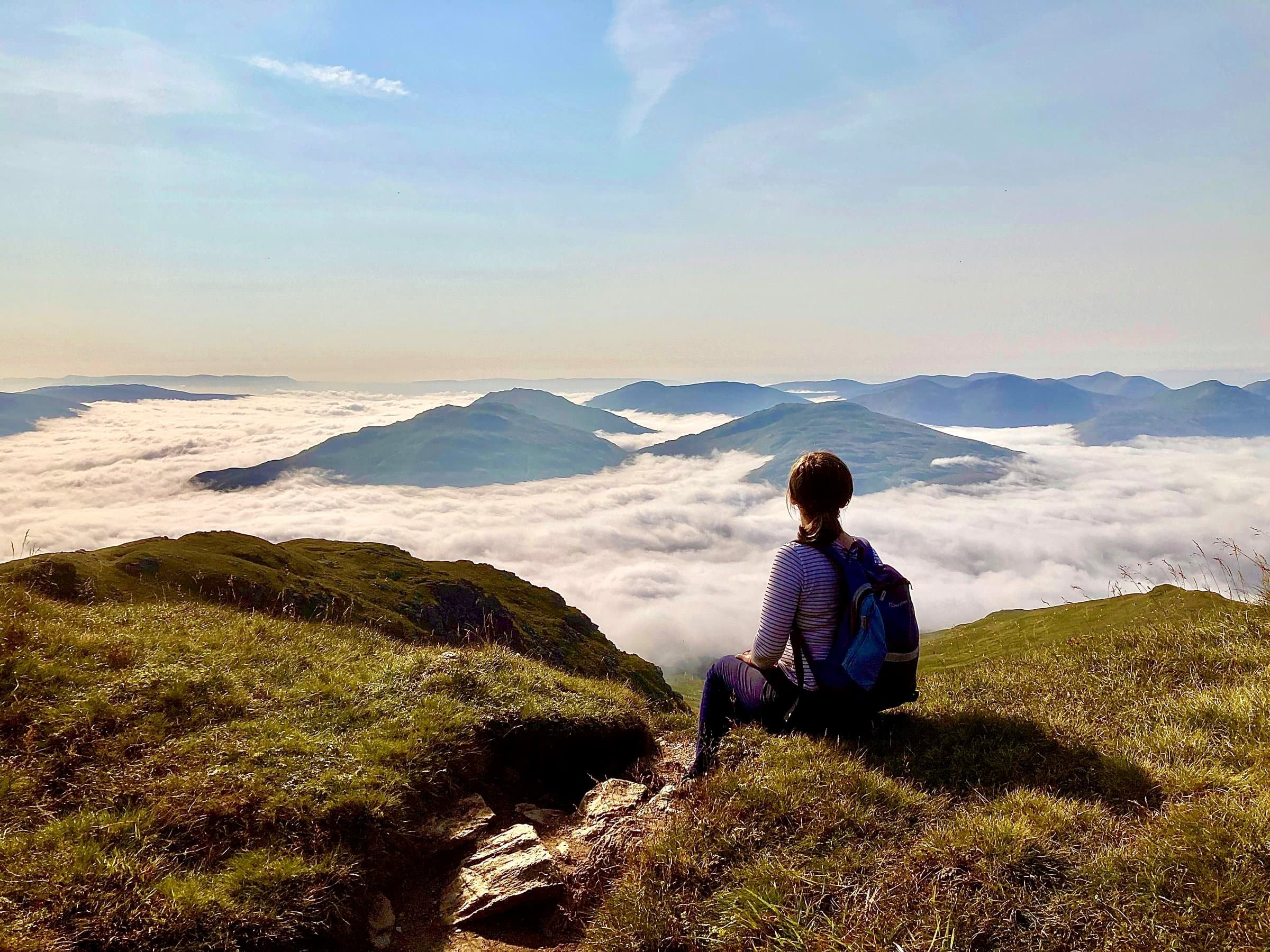 A person with a blue backpack, seen from behind, sitting on a grassy ridge above a cloud-filled valley