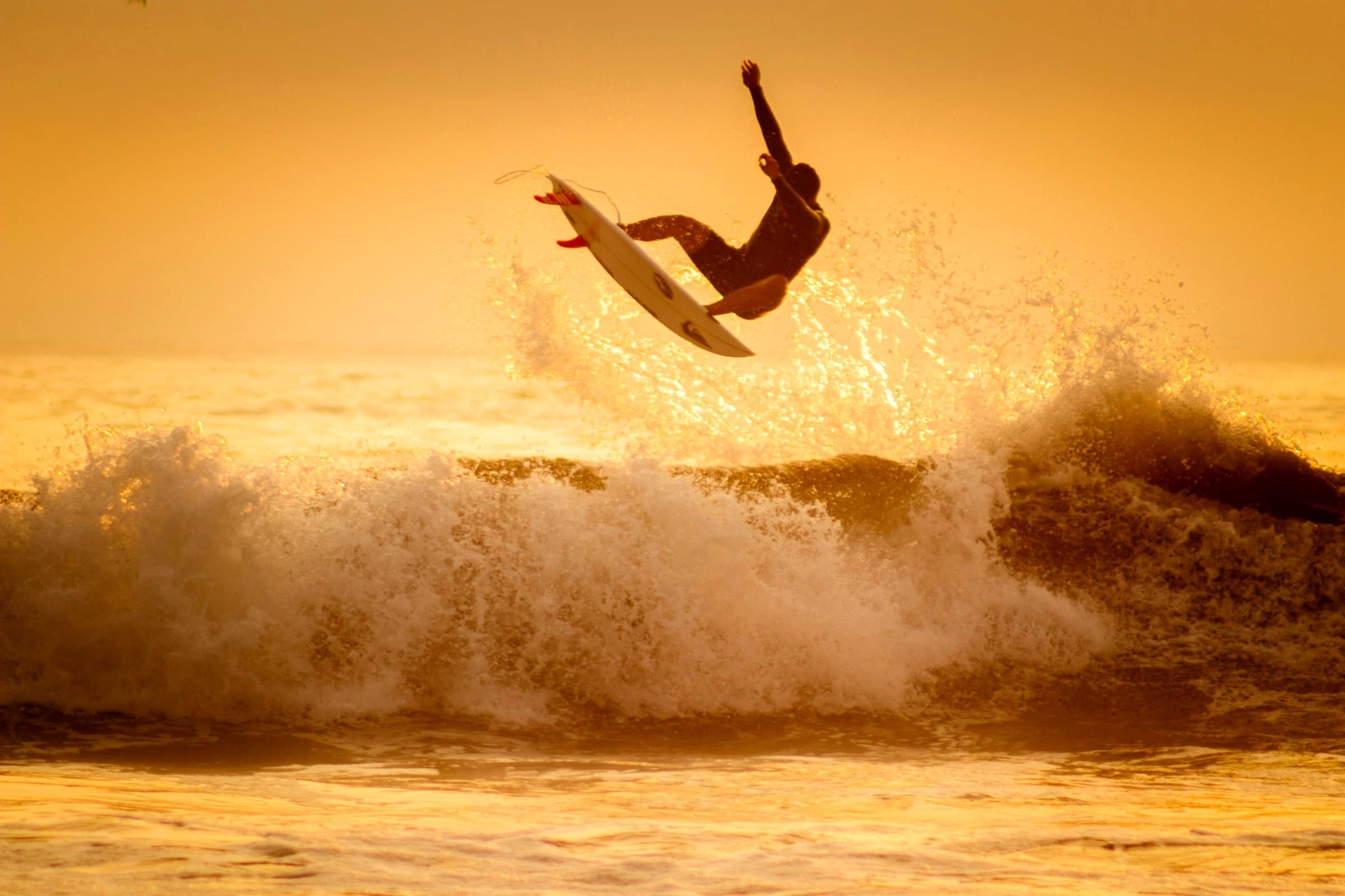 A person in a dark wetsuit holds a white surfboard in the air above a breaking wave, in golden spray